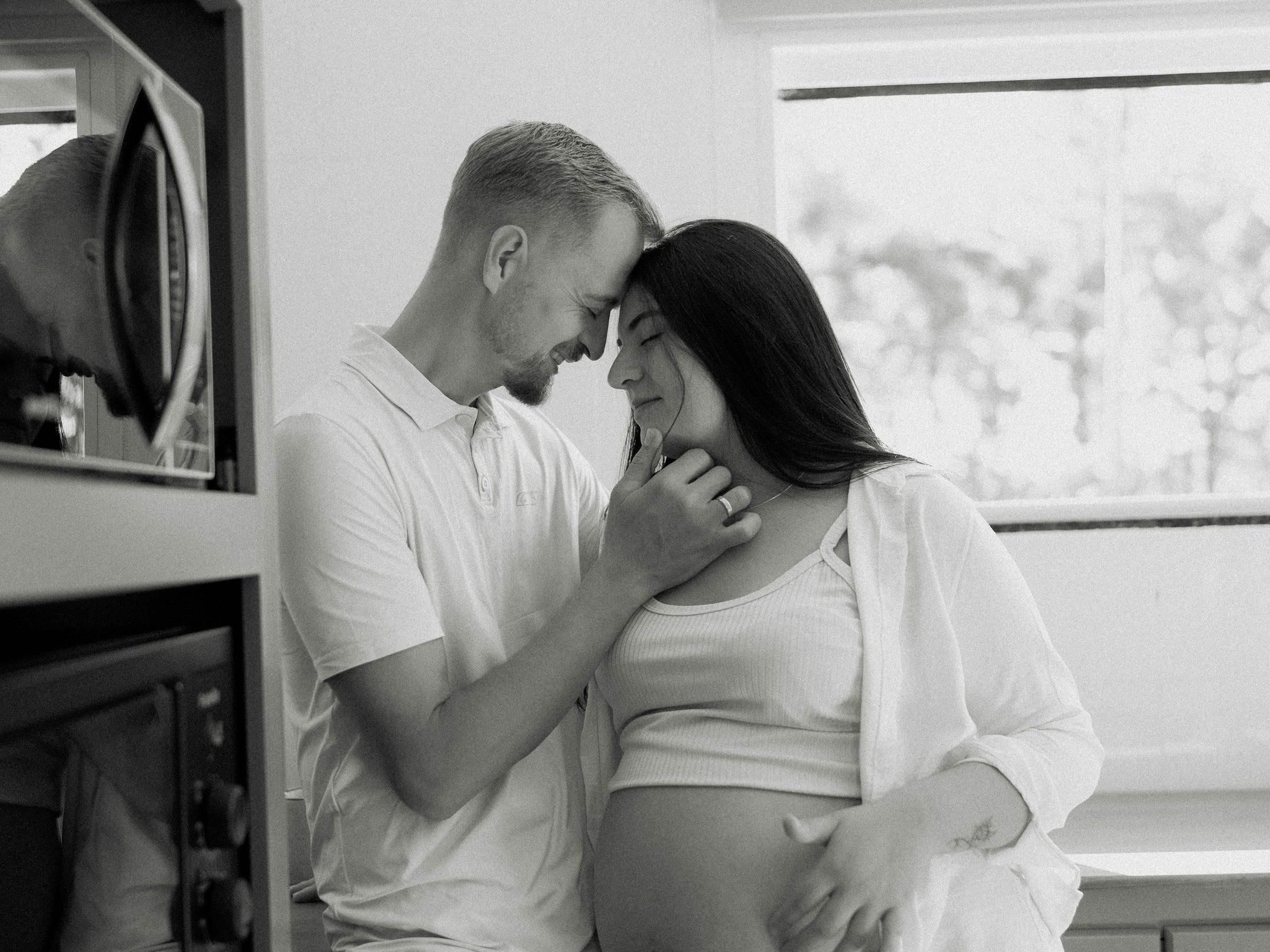 A couple standing close together in their kitchen, foreheads touching, hands on her pregnant belly